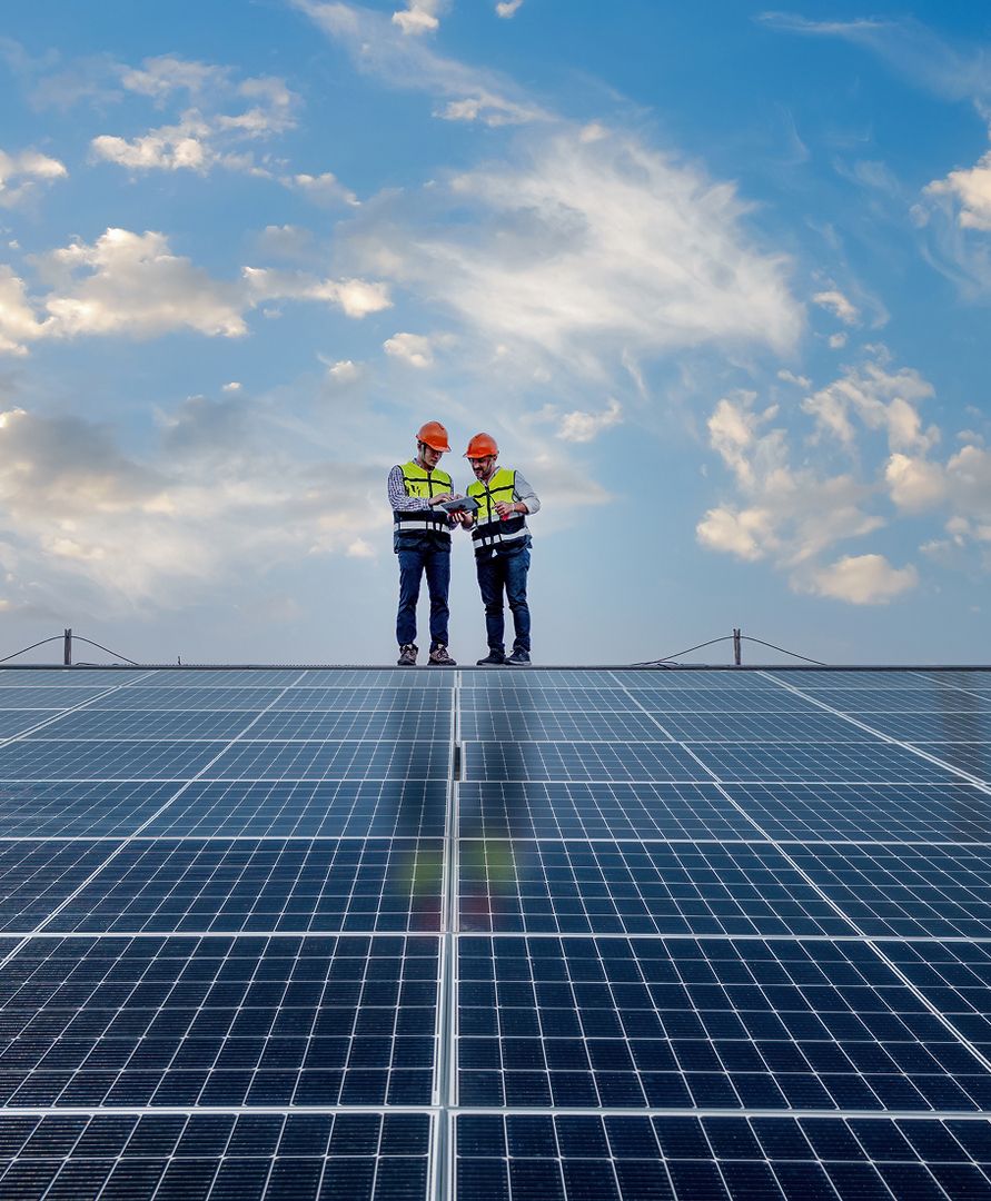 Zwei Arbeiter in Schutzkleidung stehen auf einem mit Solarzellen bedeckten Dach unter einem blauen Himmel mit vereinzelten Wolken.