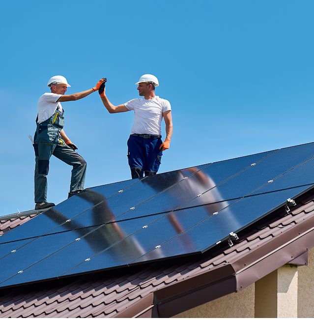 Zwei Arbeiter mit Helmen und Handschuhen bei Wartungsarbeiten auf einem mit Solarzellen bedeckten Dach unter strahlend blauem Himmel.